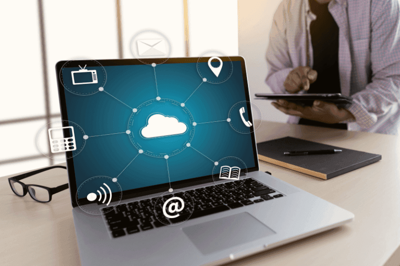 Laptop showing a cloud linked to communication icons, with a person using a tablet beside it